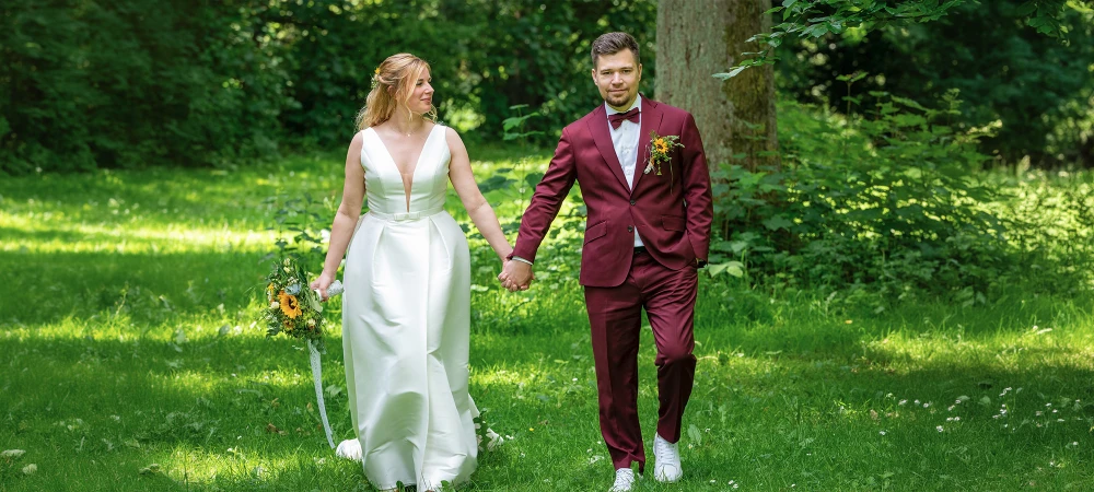 Hochzeitspaar draußen im Grünen halten Händchen und laufen auf die Kamera zu.